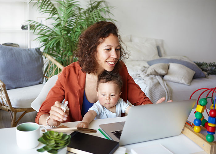 Image of woman working on laptop, holding a baby